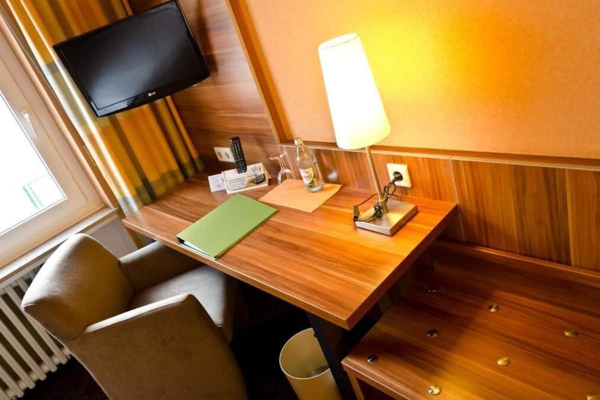 A hotel room with a wooden desk, a television mounted on the wall, a lamp, and a chair.