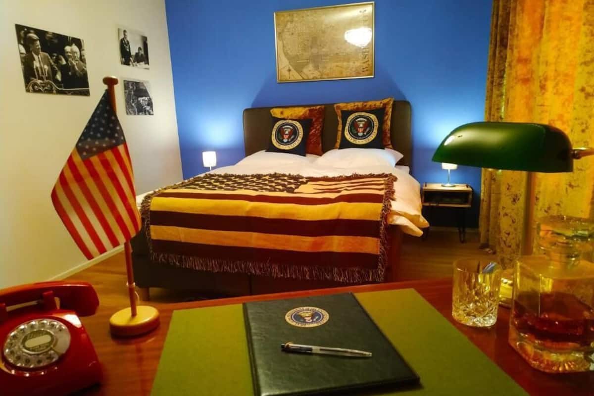 A hotel room with a bed, a desk with a telephone, a lamp, and a glass of water.