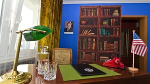 A desk with a green desk lamp, a laptop, a red rotary phone, and a framed picture. There is a bookshelf with books and an American flag. - Hotel Rheinland (Bonn)