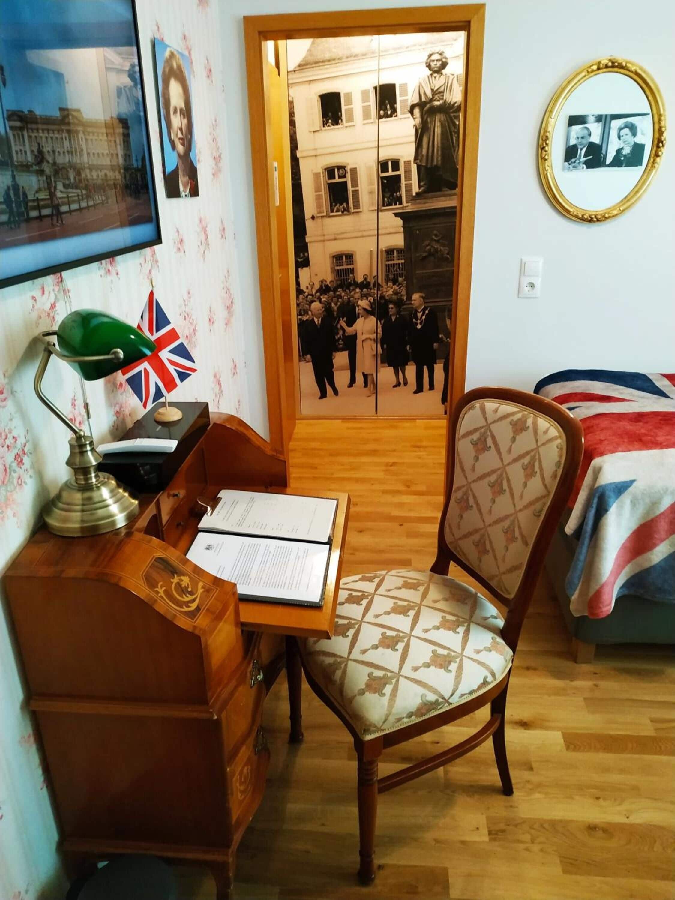 A room with a wooden desk, a chair, a bed with a Union Jack blanket, and a mirror on the wall.