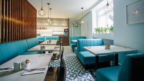 A modern restaurant with blue booths and tables, wooden slats on the left wall, and a kitchen in the background. - Mentone Hotel (London)