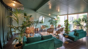 A spacious living room with a green sofa, wicker chairs, and a dining area. There are multiple potted plants and a large window providing natural light. - Condotti Palace (Rome)