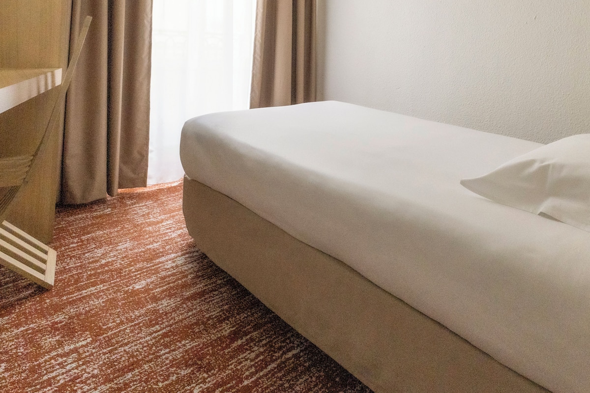 Una habitación de hotel con una cama individual, cortinas color beige y piso alfombrado.