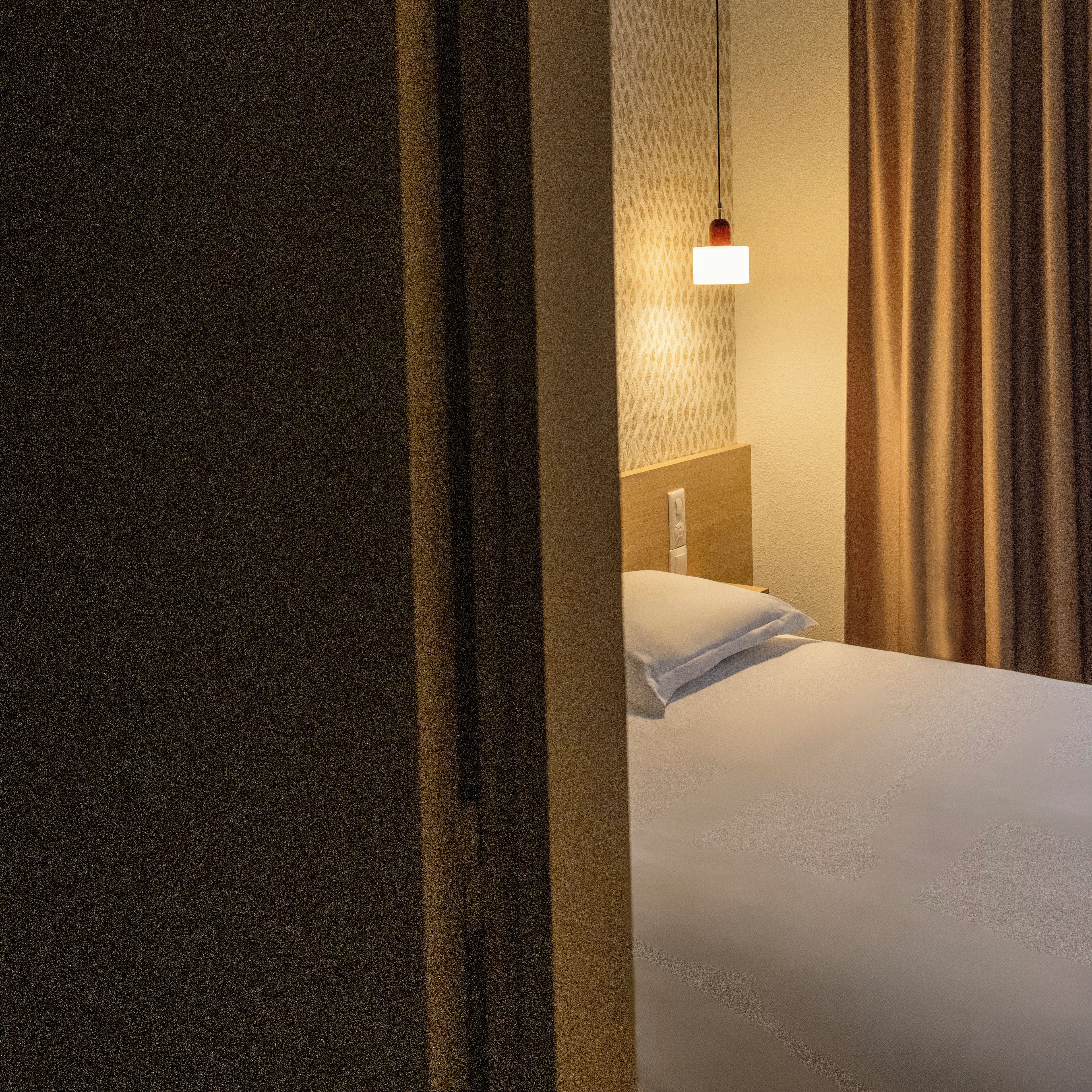 Habitación de hotel con cama, mesita de noche, lámpara de pared y ventana con cortinas.