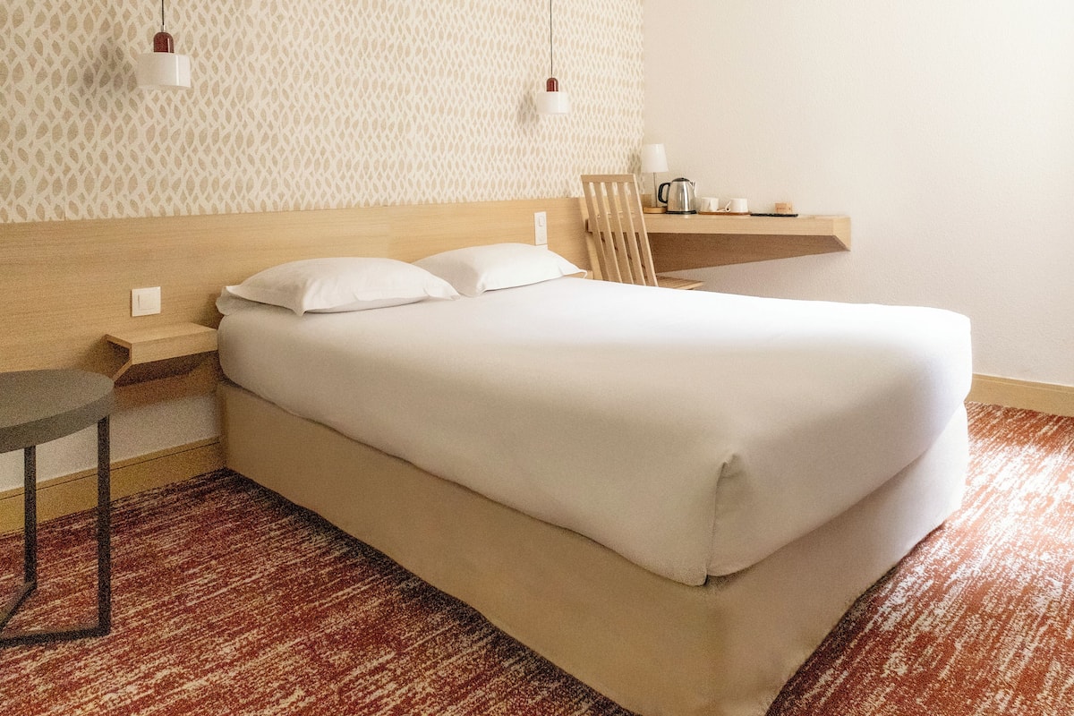 Una cama bien hecha con sábanas blancas, un cabecero de madera, una mesita de noche pequeña con una lámpara y un papel tapiz estampado.
