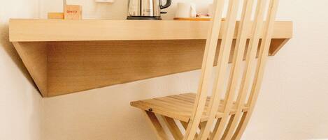 A compact room with a wooden desk, a chair, a kettle, a lamp, and a small shelf with a box labeled "GINTO".