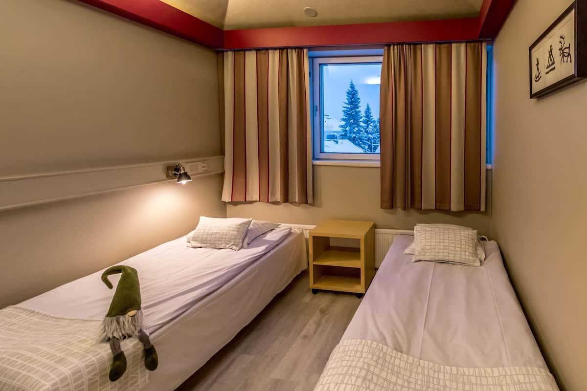 A room with two beds, a window with curtains, a nightstand, and a wall-mounted lamp.