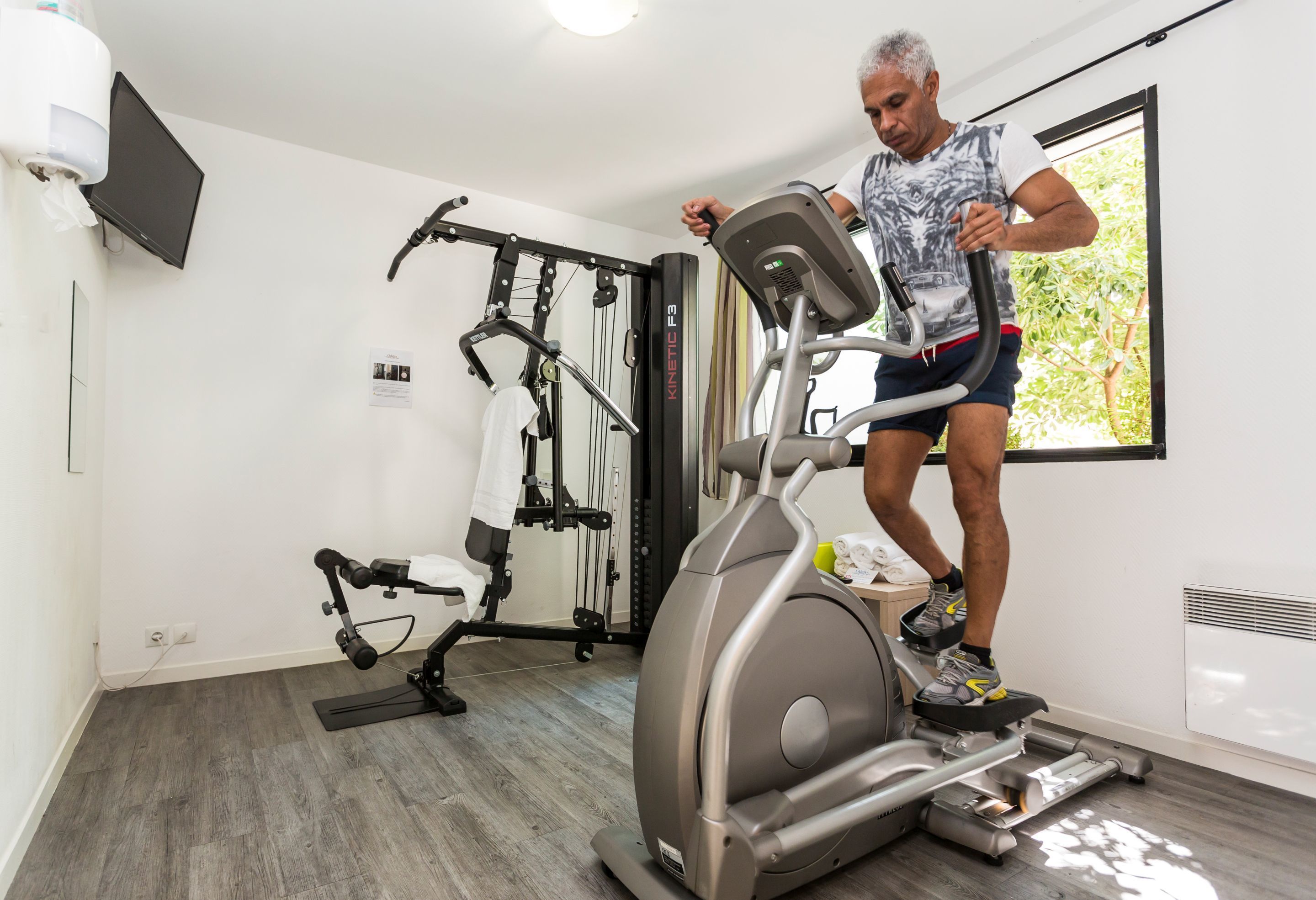 A man using an elliptical machine in a gym with a television mounted on the wall and a window with a view of trees.