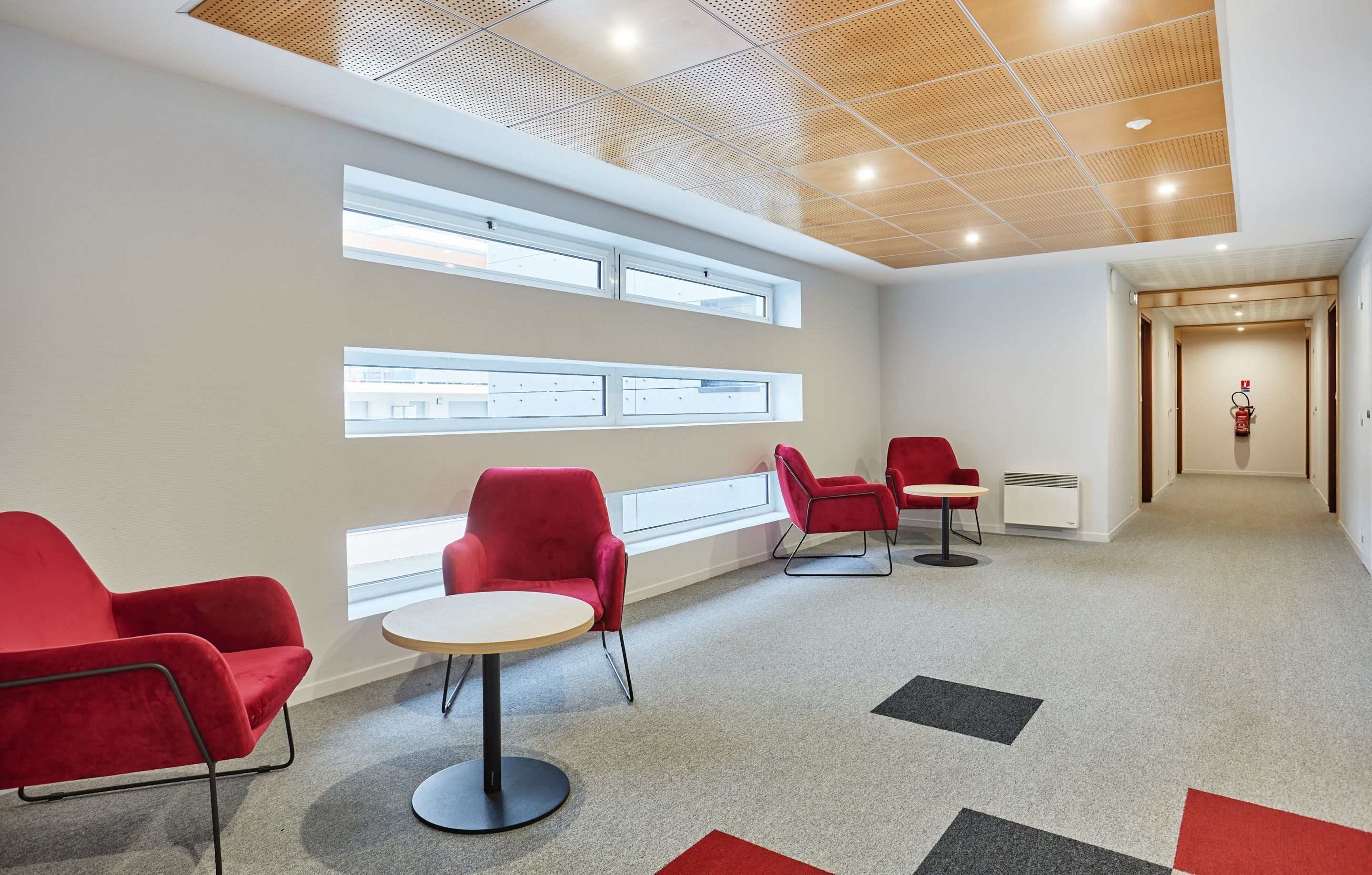 Un área de espera moderna con sillas rojas, una mesa redonda y ventanas amplias que dejan entrar luz natural.