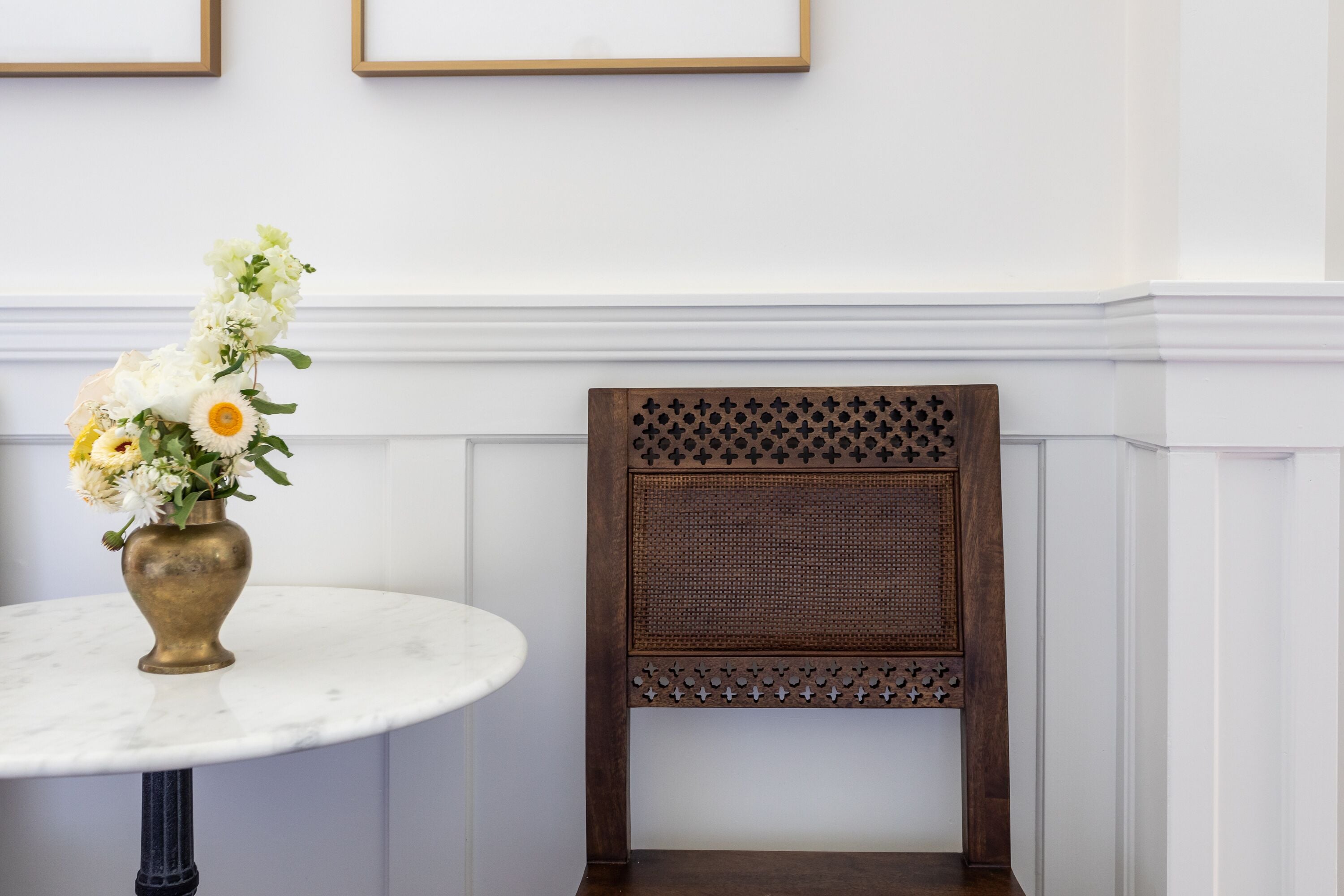 Une table en marbre blanc surmontée d’un vase en laiton rempli de fleurs, une chaise en bois avec un dossier ajouré et un mur blanc orné de deux tableaux encadrés.