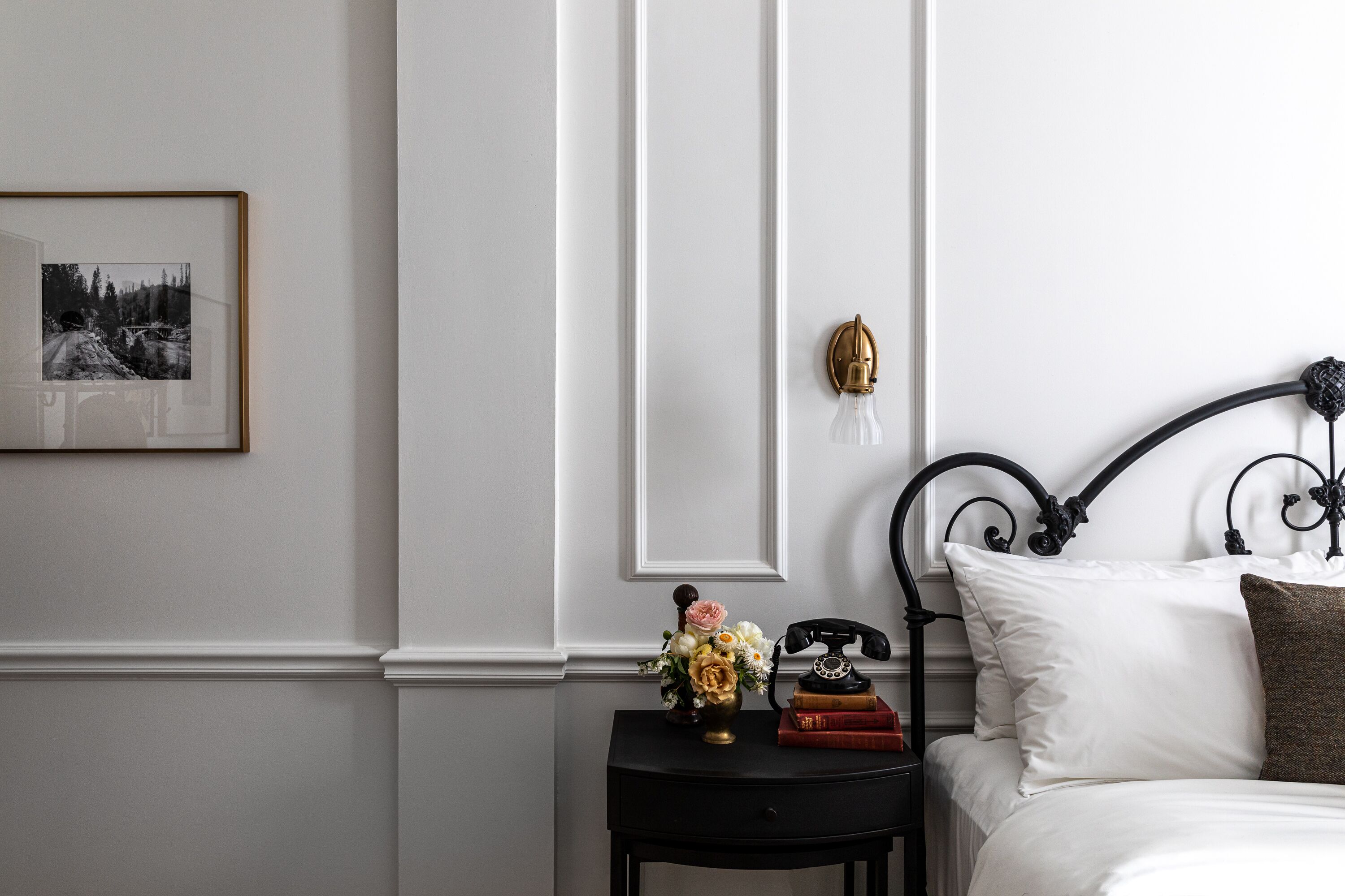 A neatly arranged bedroom with a bed, a bedside table with a telephone, a framed picture, and a vase with flowers.
