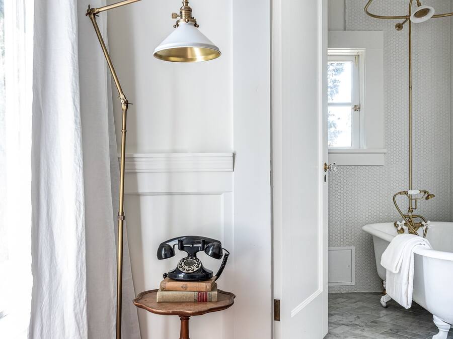 A room with a wooden floor, a vintage telephone on a small table, a brass floor lamp, and a white bathtub.