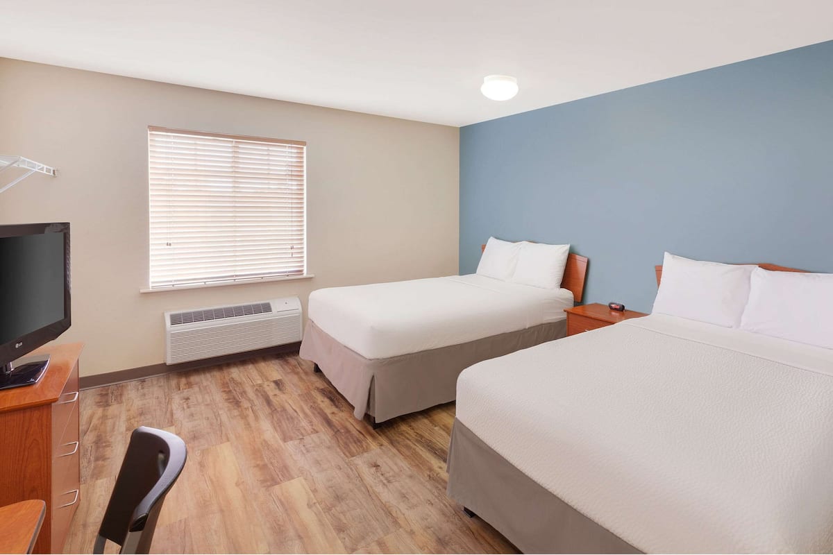 Une chambre d’hôtel avec deux lits, un bureau avec une télévision, une chaise, une fenêtre avec des stores et un radiateur.