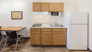A compact kitchen with wooden cabinets, a microwave, a refrigerator, a sink, and a dining table with chairs. - WoodSpring Suites Des Moines Pleasant Hill (Pleasant Hill)