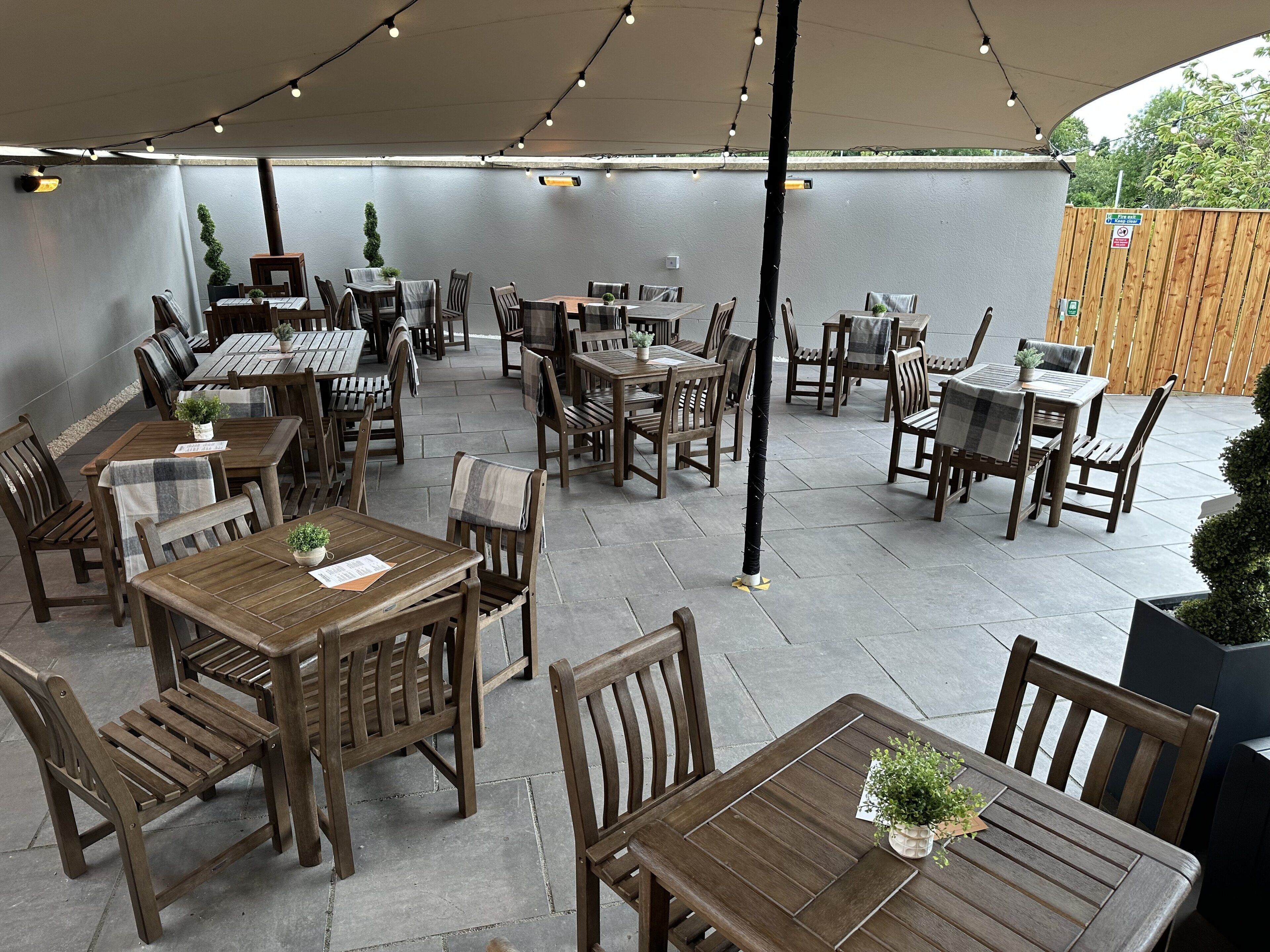 An outdoor patio with multiple wooden tables and chairs, string lights overhead, and potted plants.