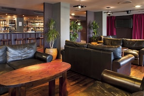 Una zona de bar con mostrador y taburetes, un área de estar con sofás de cuero y una mesa de madera, y un televisor en la pared.