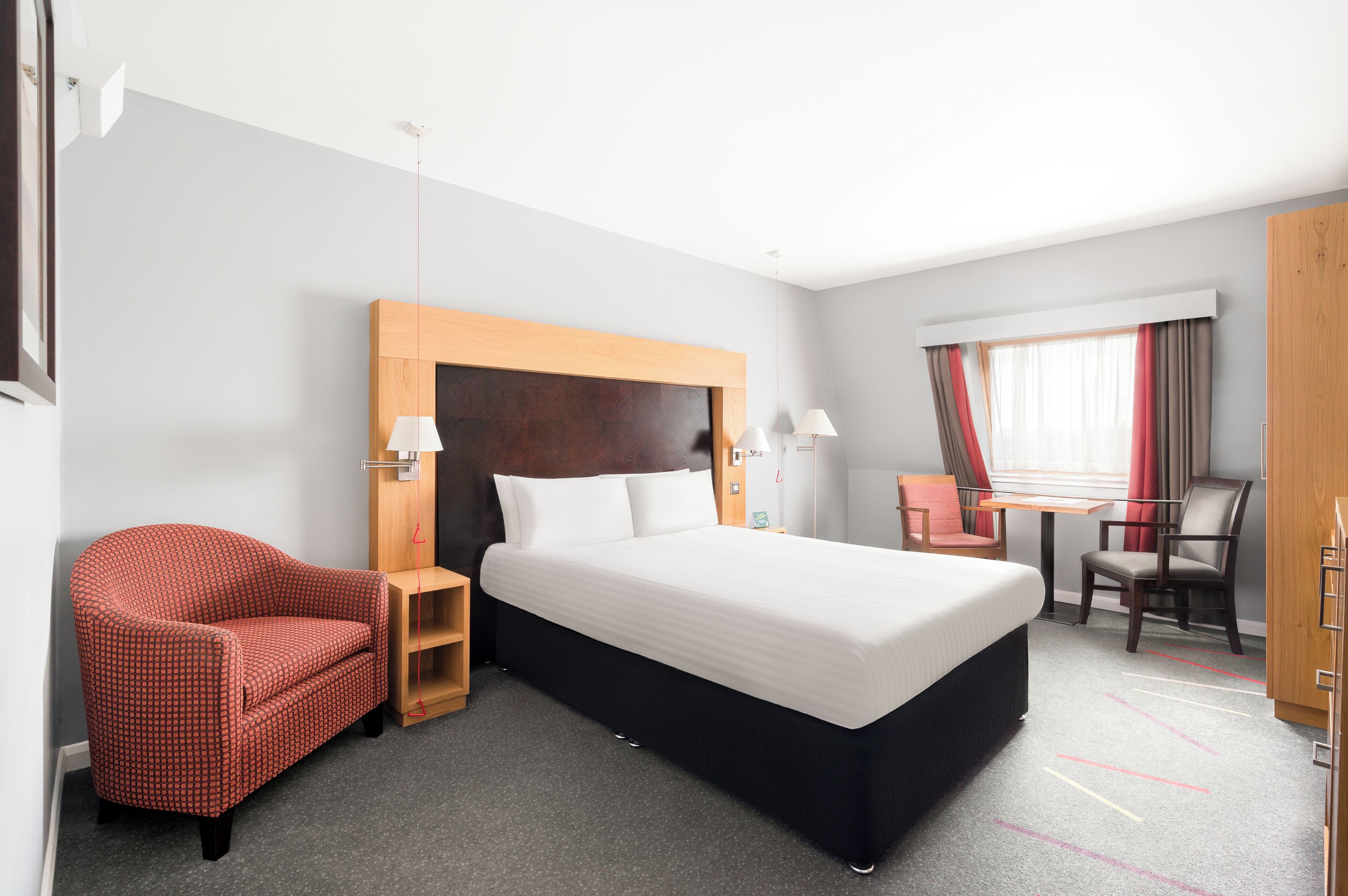 Habitación de hotel con una cama grande, un sillón rojo, una mesita de noche de madera, un escritorio con silla y un armario.