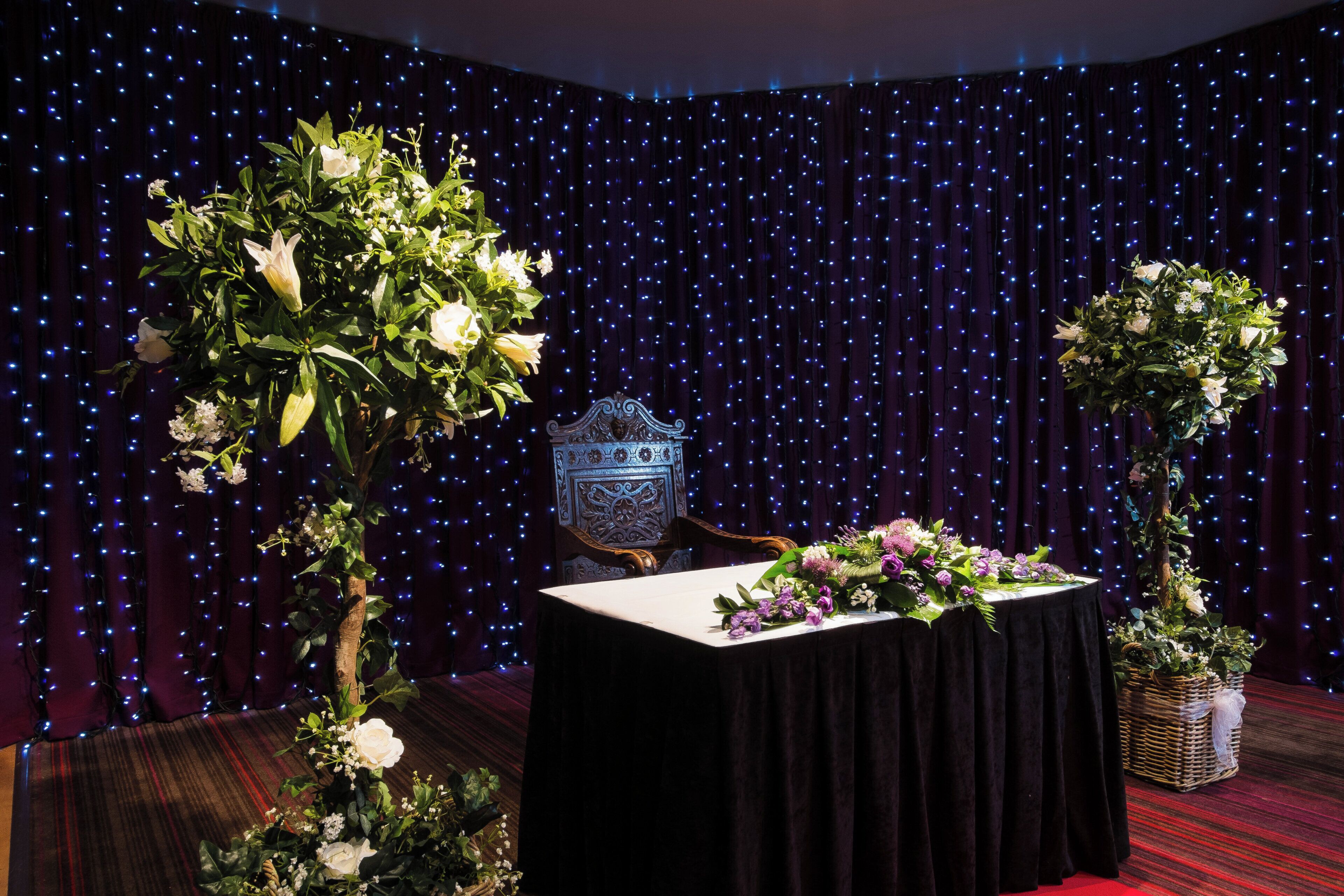 Ett rum med en mörk draperad bakgrund, ett bord med ett vitt bordslinne och blomsterarrangemang.