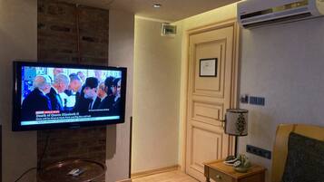 Een hotelkamer met een televisie waarop een nieuwsbulletin draait, een klein rond tafeltje, een kamerbreed tapijt met dessin, een houten nachtkastje en een bed met een beddegoed met patroon.