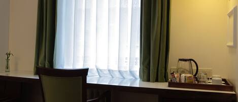 A hotel room with a wooden desk, a chair, a window with sheer curtains, a wall-mounted light fixture, and a framed picture of wine bottles.