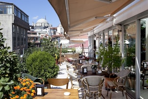 An outdoor cafe with wicker furniture, potted plants, and a view of historic buildings in the background.