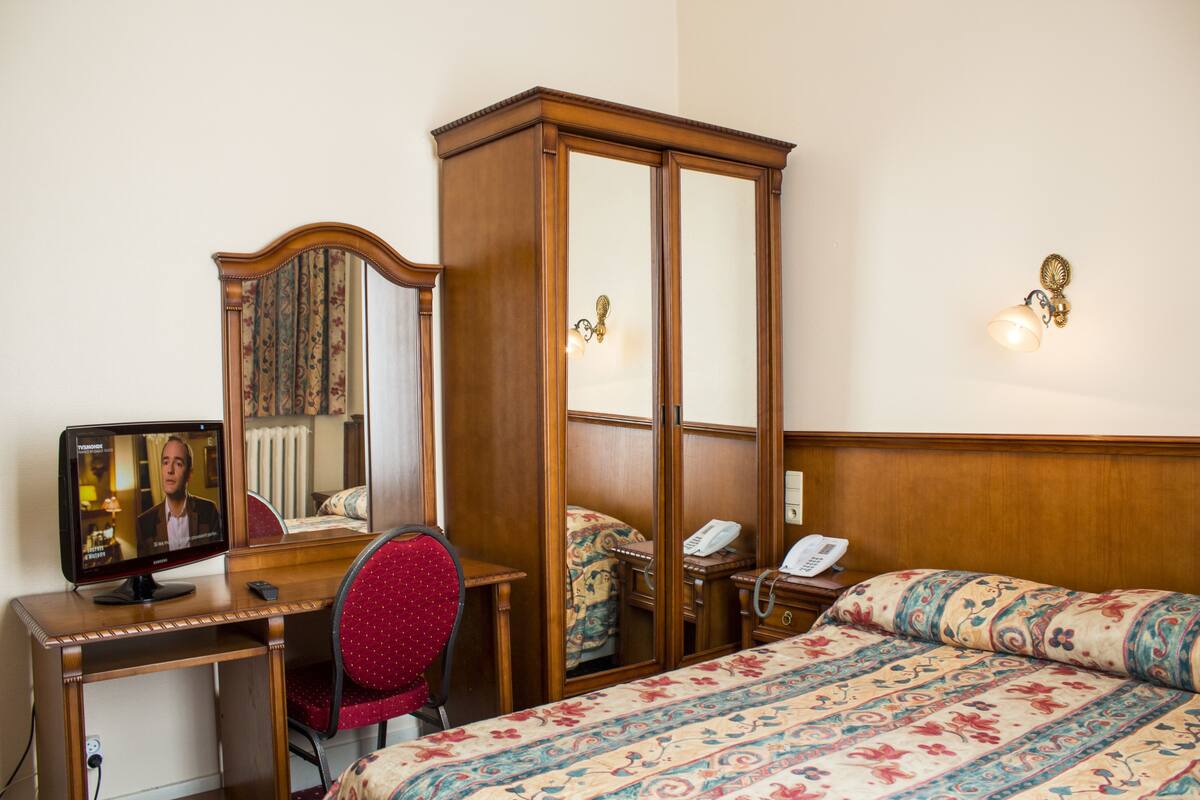 Ein Hotelzimmer mit einem Holztisch, einem roten Sessel, einem Bett mit gemusterten Betttextilien, einem Kleiderschrank und einem Fernseher, der einen Mann zeigt.