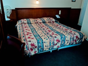 Una cama individual con una colcha de motivos florales en una habitación con cabecera de madera y mesita de noche con lámpara.