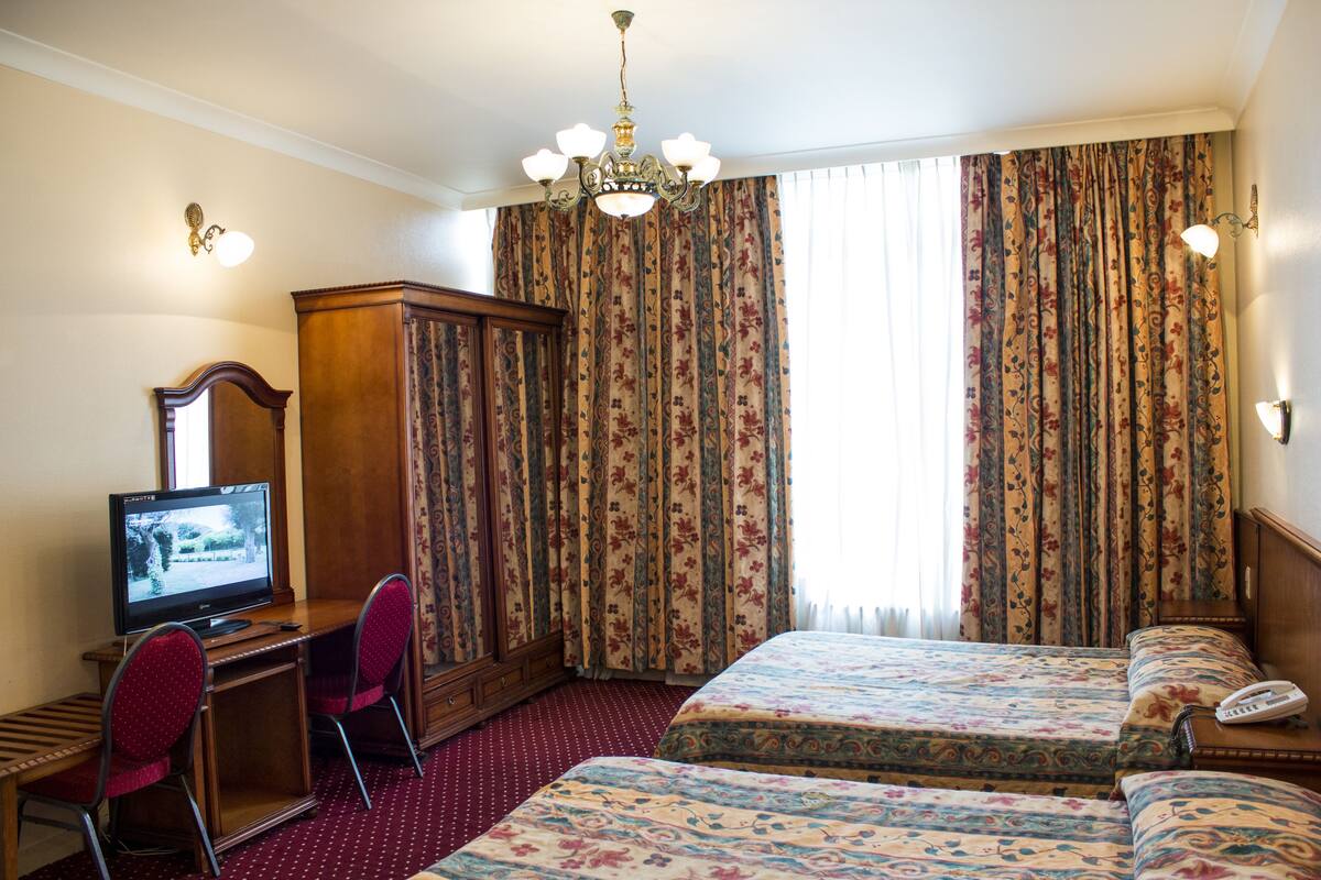 Ein Hotelzimmer mit Schreibtisch aus Holz, zwei roten Sesseln, einem Fernseher, einem Kleiderschrank, einem Bett mit floralem Bettbezug und einer Kristallleuchter.