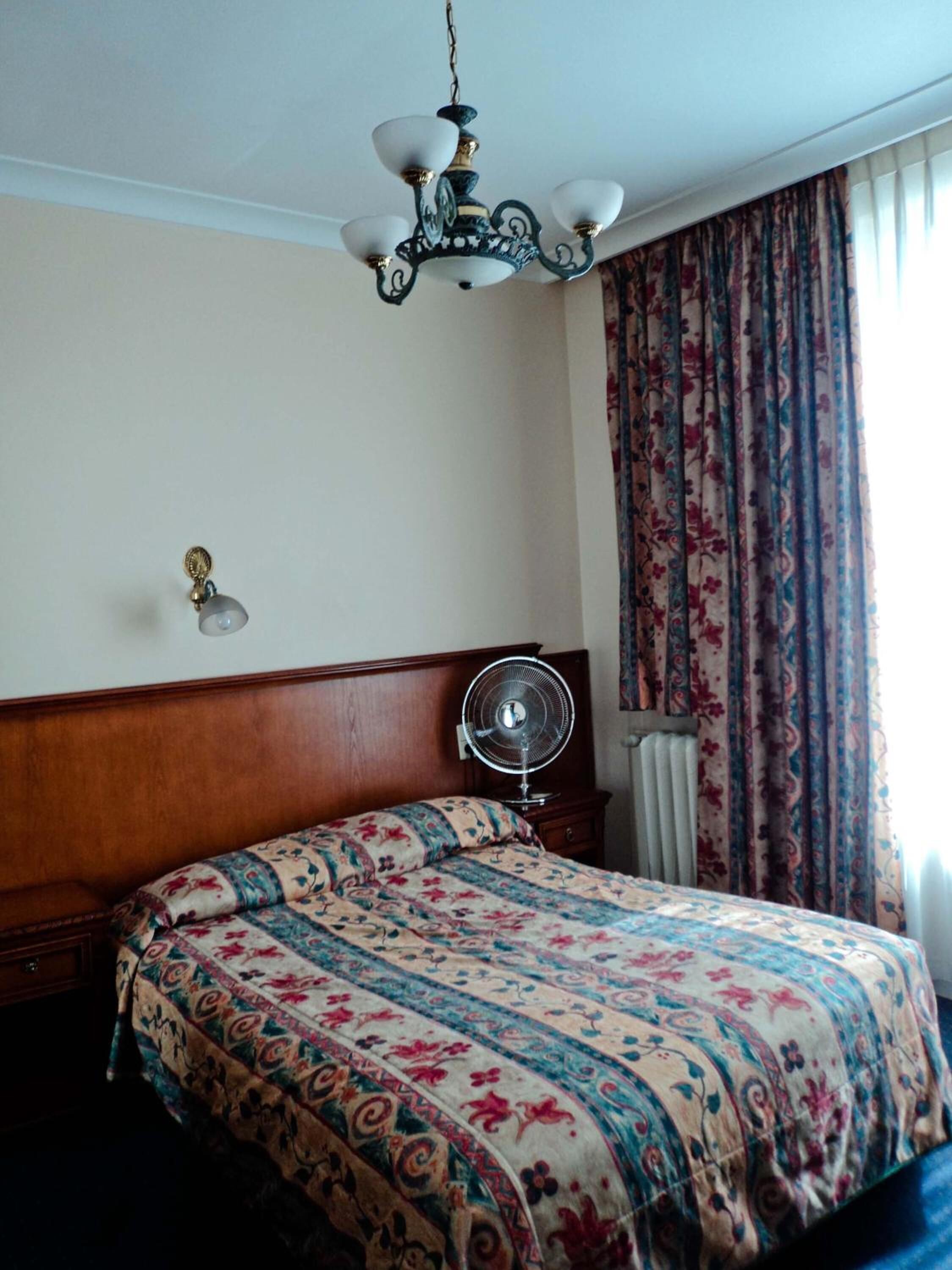Una habitación de hotel con cabecera de madera, cama con colcha de estampado floral, ventilador de techo y ventana con cortinas estampadas.