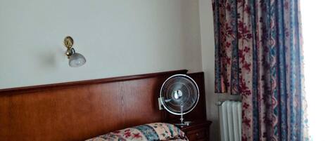 Una habitación de hotel con cabecera de madera, cama con colcha de estampado floral, ventilador de techo y ventana con cortinas estampadas.