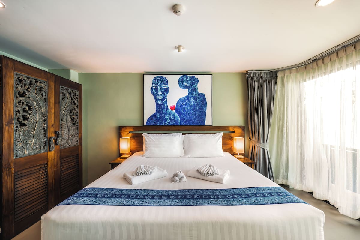 A neatly made bed with white linens and a blue accent, a large painting of two figures above it, and a wooden wardrobe with intricate carvings to the left.
