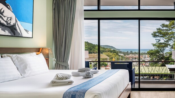 A hotel room with a large bed, neatly folded towels, a view of the city and hills through large windows, and a painting on the wall.