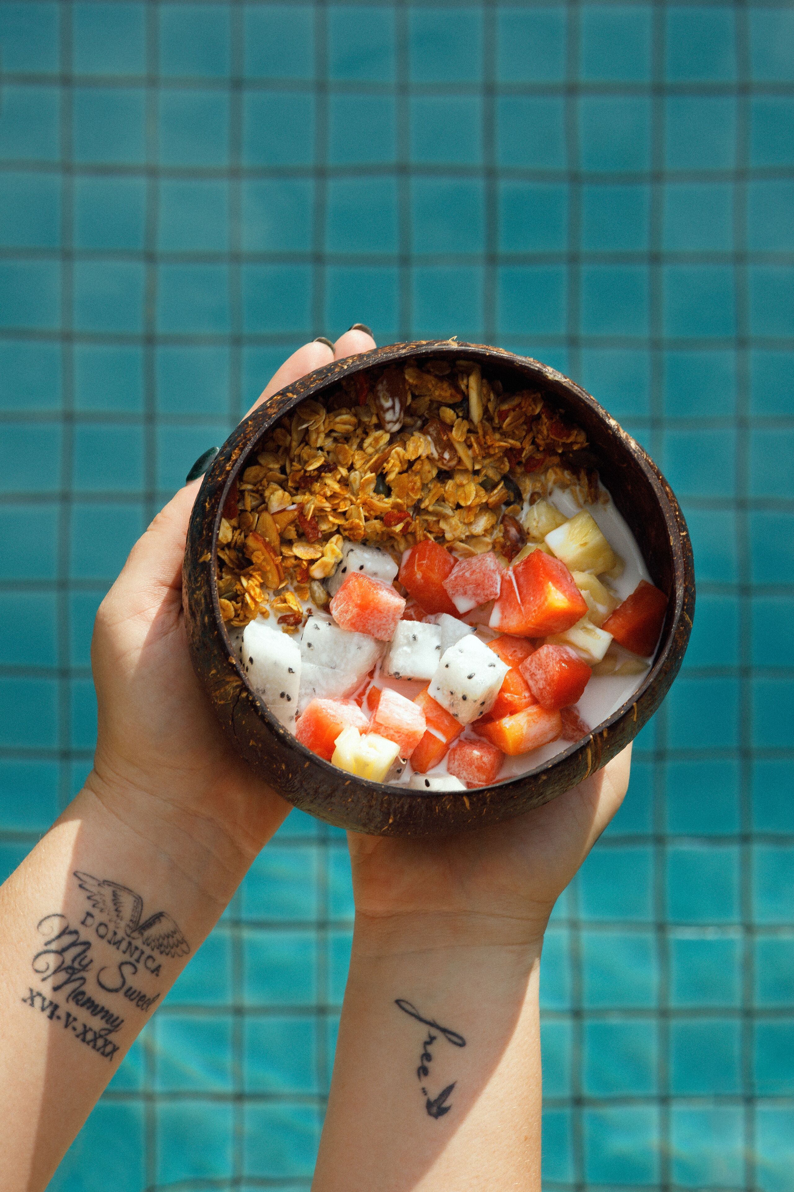 Una mano sosteniendo un tazón con fruta y granola junto a una piscina.
