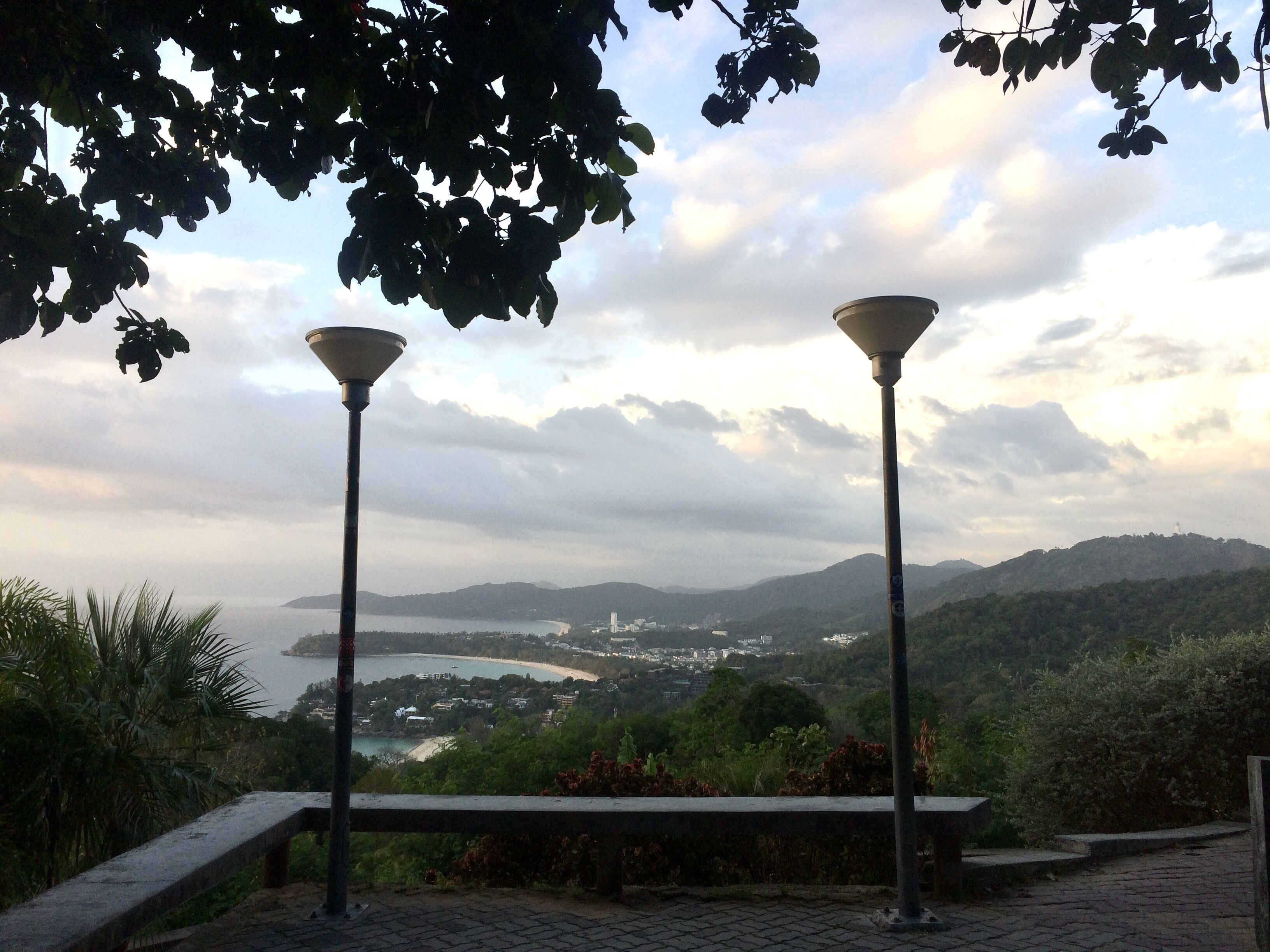 A bench with two street lamps, overlooking a coastal landscape with hills and a body of water.