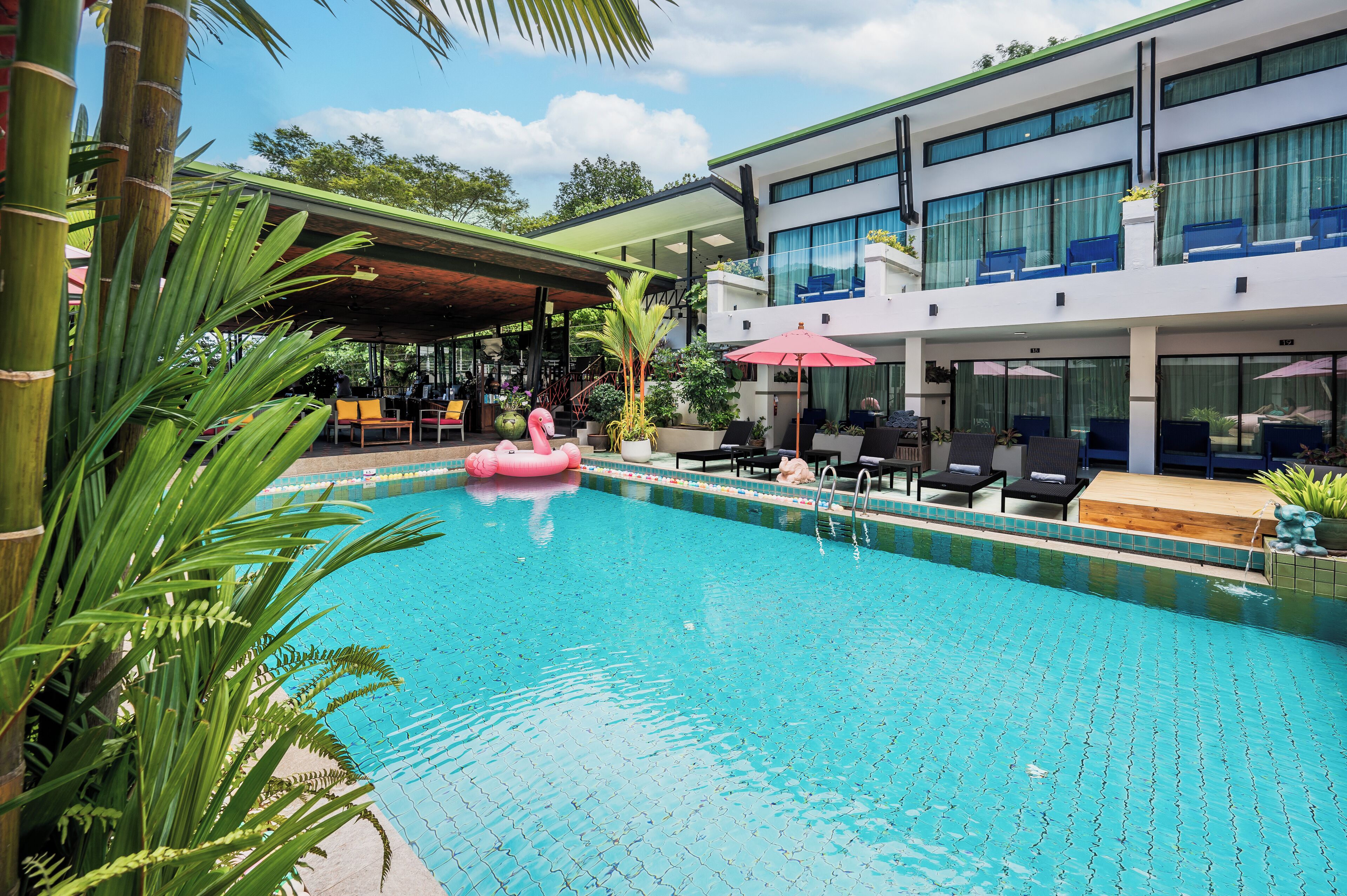 A swimming pool with lounge chairs and umbrellas, surrounded by greenery and a modern building with large windows.