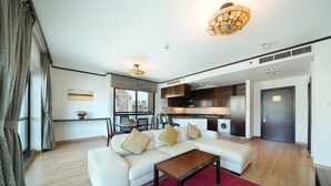 Un salon moderne avec un canapé blanc, une table basse en bois et un tapis rouge. On y trouve également un coin cuisine avec des meubles sombres et un réfrigérateur en acier inoxydable. Un coin repas, avec une table et des chaises, est visible au fond.