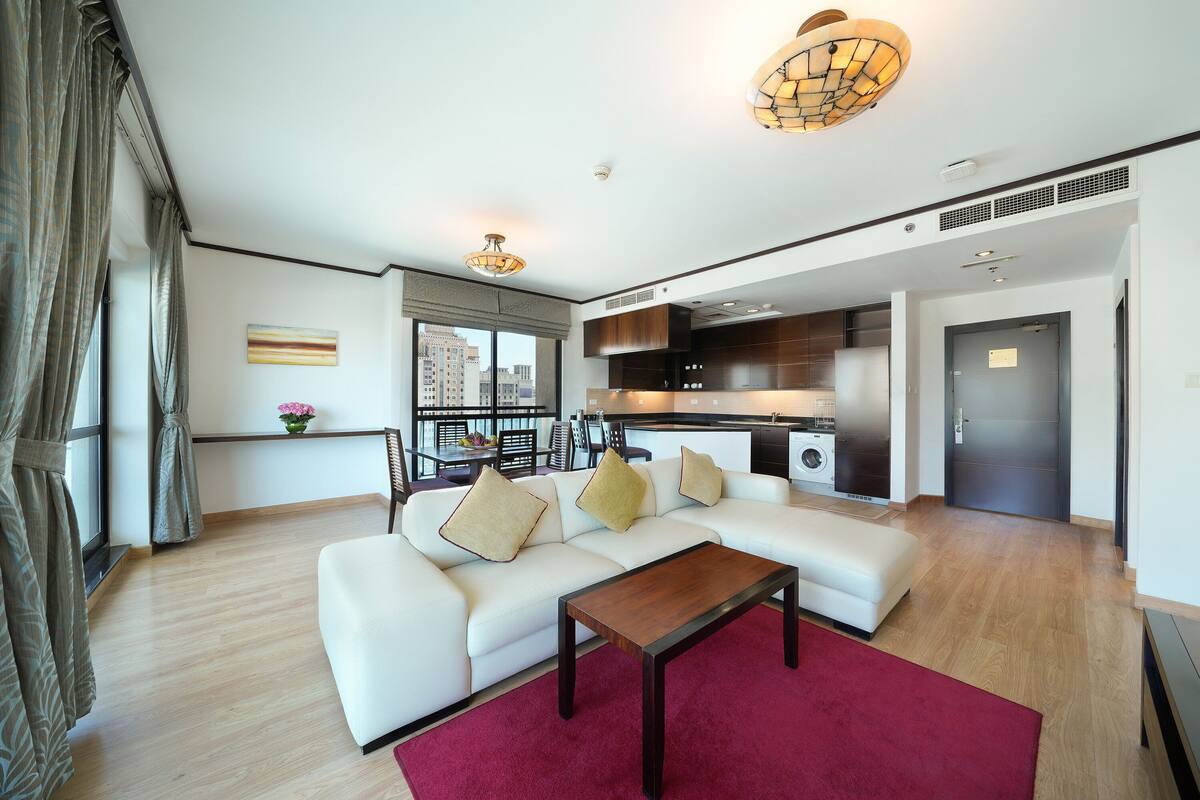 Ein modernes Wohnzimmer mit einem weißen Sofa, einem Holztisch und einem roten Teppich. Es gibt eine Küchenzeile mit dunklen Schränken und einem Edelstahlkühlschrank. Im Hintergrund ist ein Essbereich mit Tisch und Stühlen zu sehen.