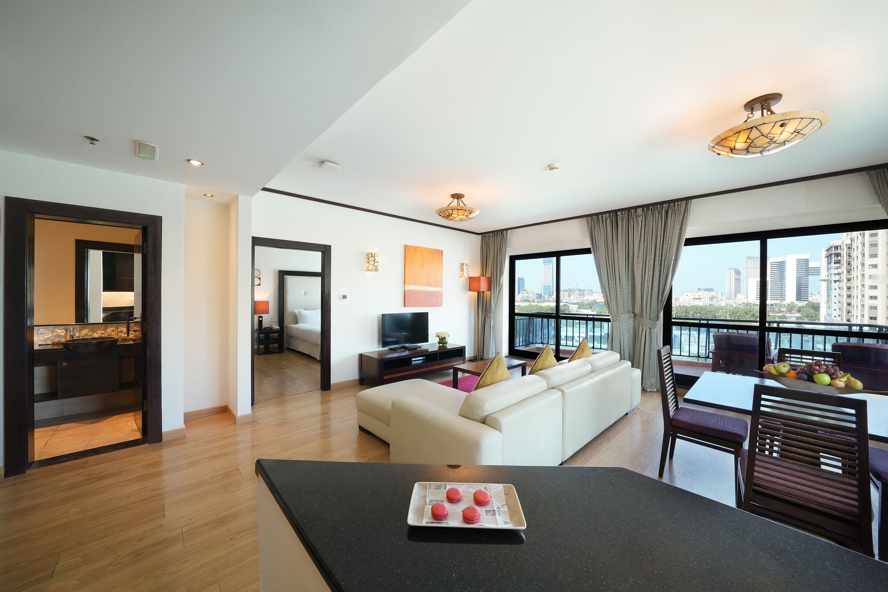 A modern living room with a sofa, dining table, and chairs. There is a TV and a small table with a fruit bowl. The room has large windows with a city view.