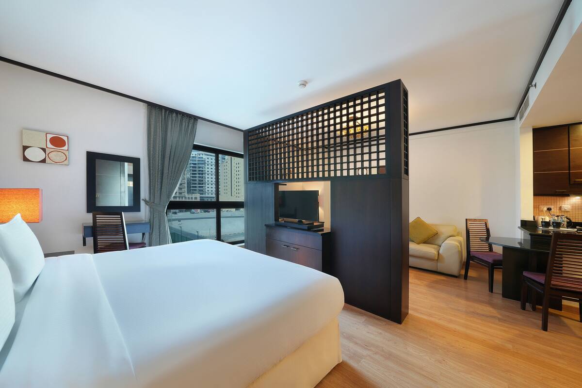 Ein Hotelzimmer mit einem großen Bett, einem Fernseher, einem Schreibtisch mit Stühlen und Blick auf die Stadt.