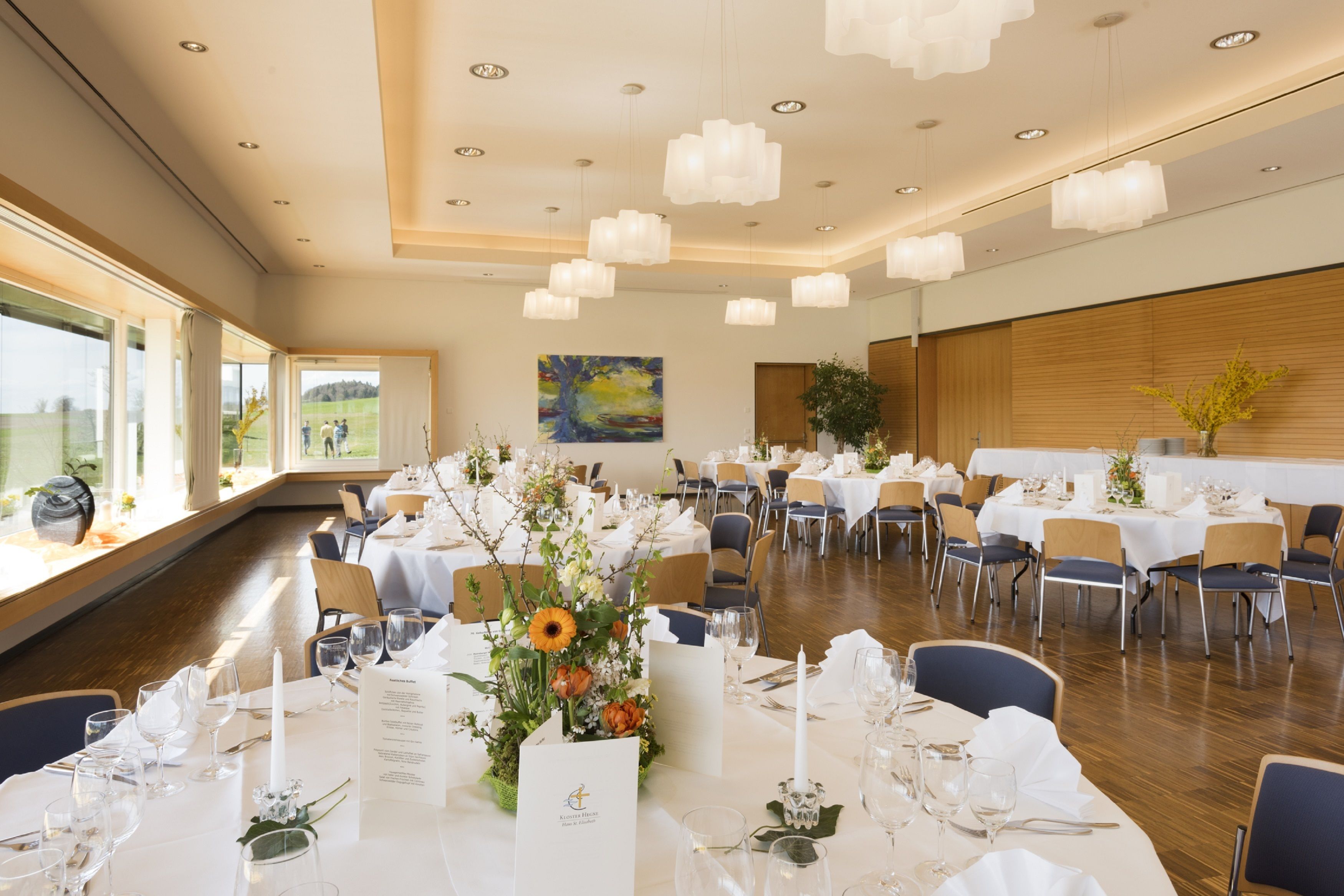 A spacious banquet hall with multiple tables set for dining, large windows offering a view of the outdoors, and a modern ceiling design with recessed lighting.