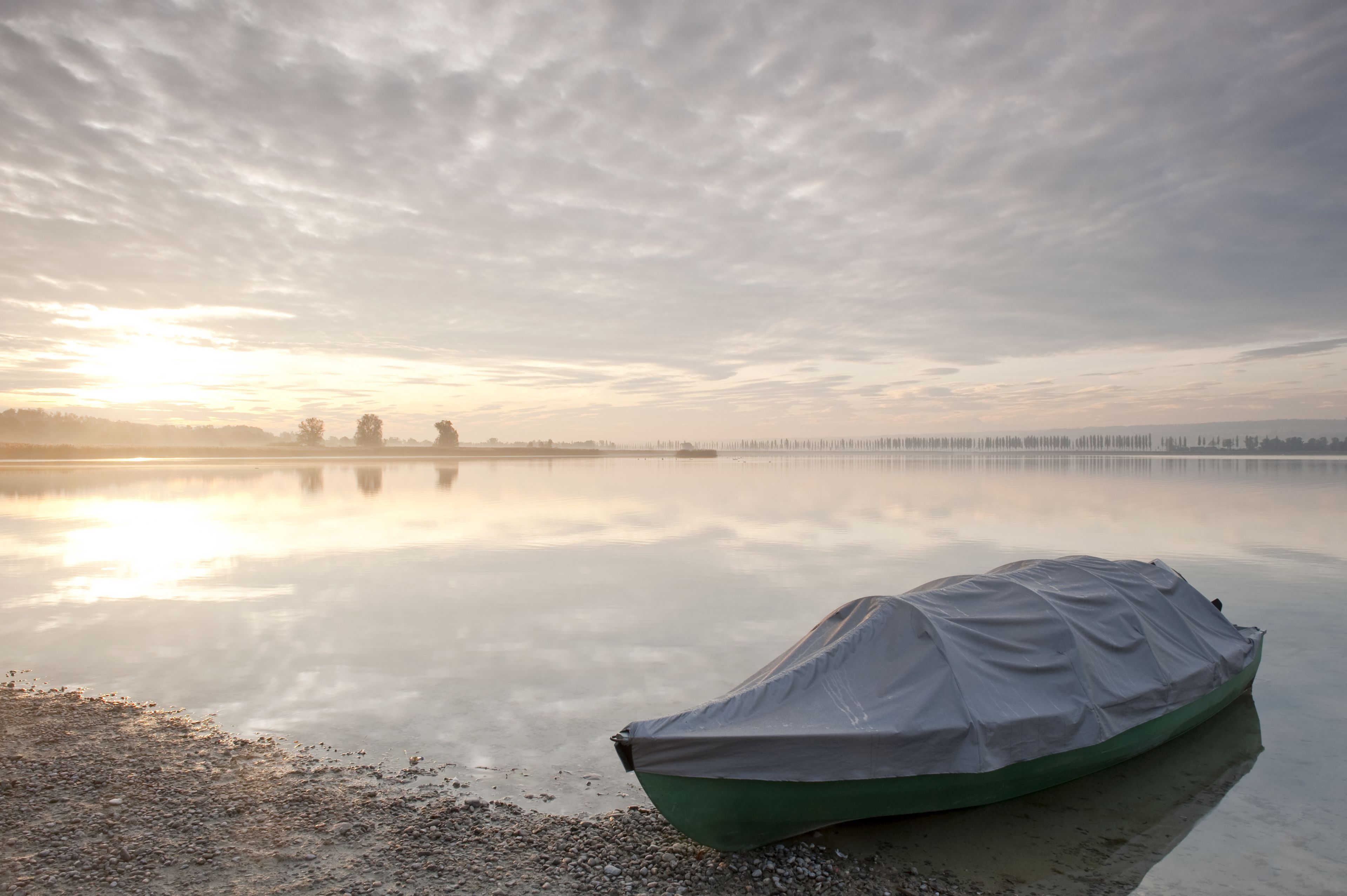 Un lago tranquilo con un bote cubierto en la orilla y una costa distante con árboles y un puente.