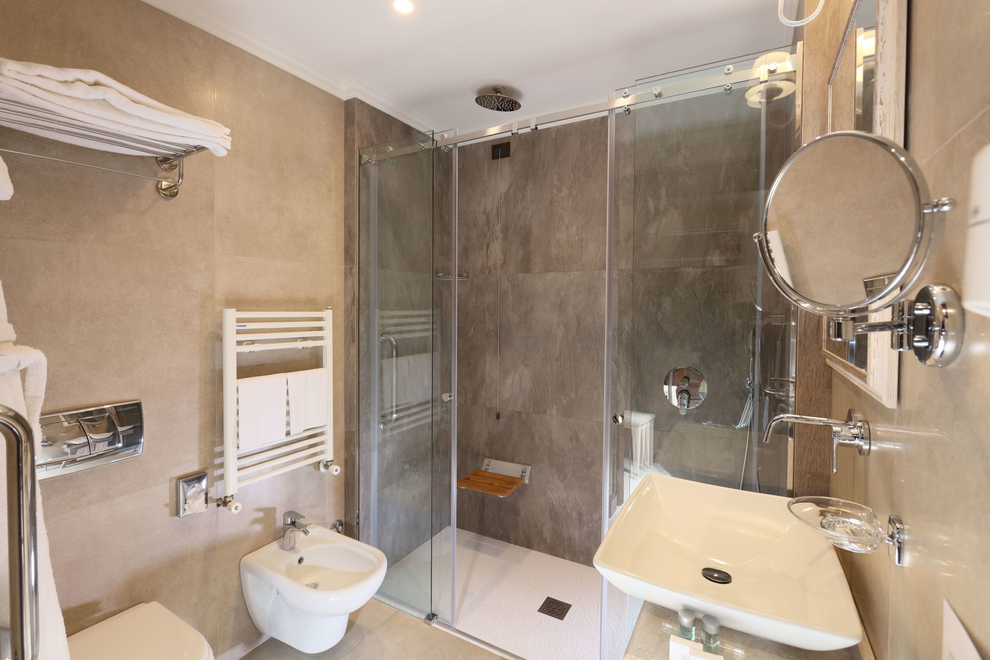 A modern bathroom with a glass-enclosed shower, a bidet, a sink with a glass soap dish, and a towel rack.