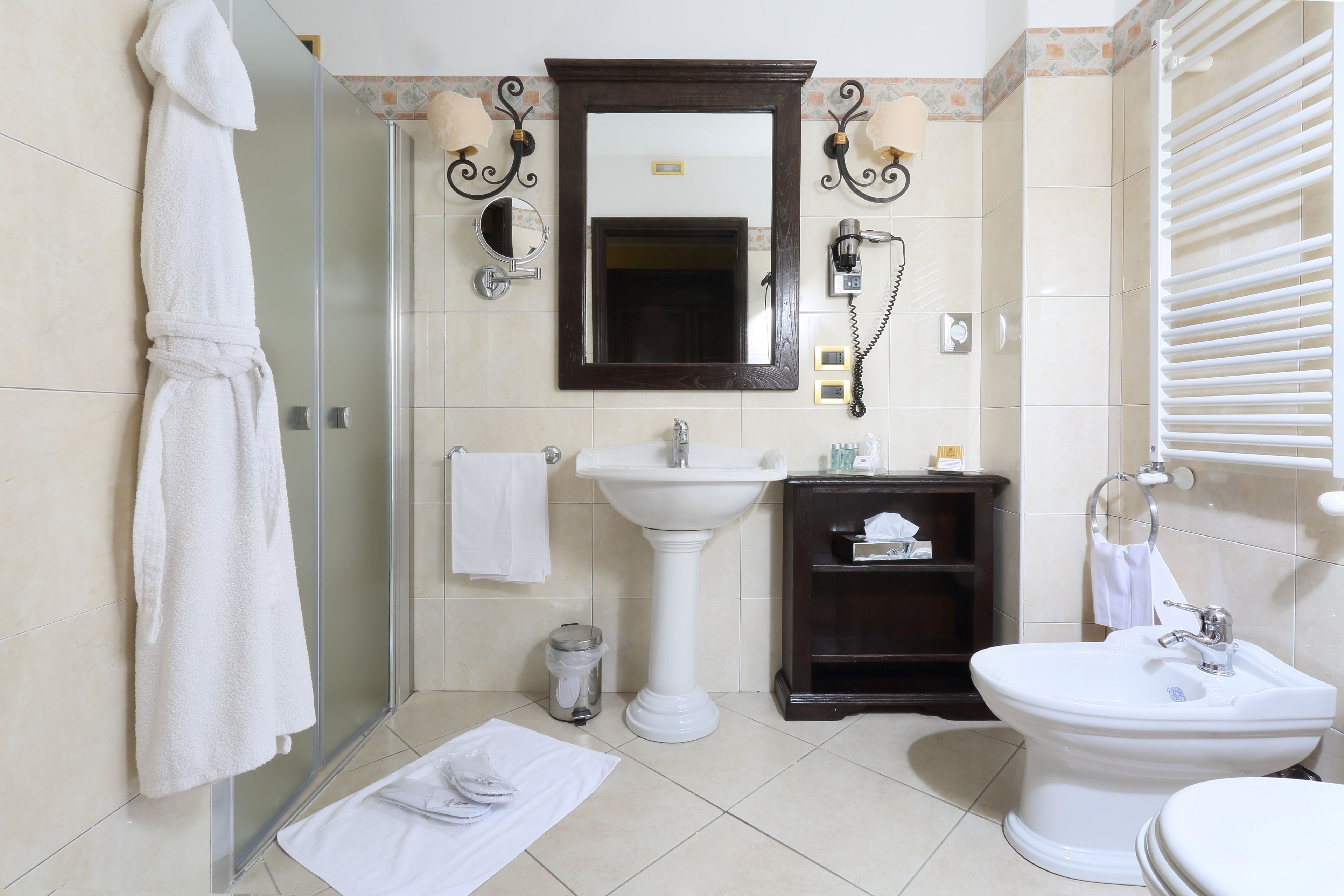 A bathroom with a glass shower enclosure, a pedestal sink, a toilet, and a vanity with a mirror and wall sconces.