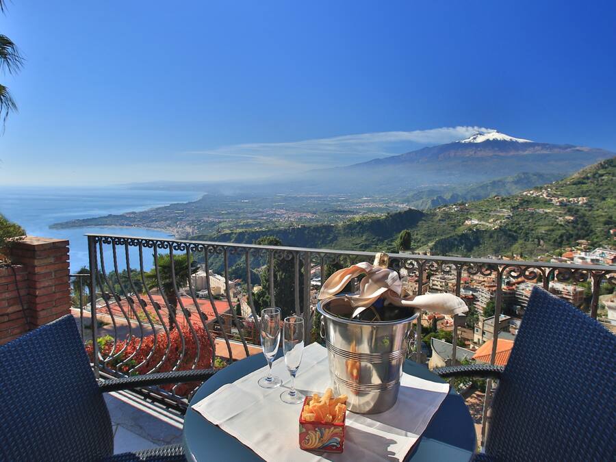 A balcony with a table set for two, overlooking a scenic view of a mountain and the sea.