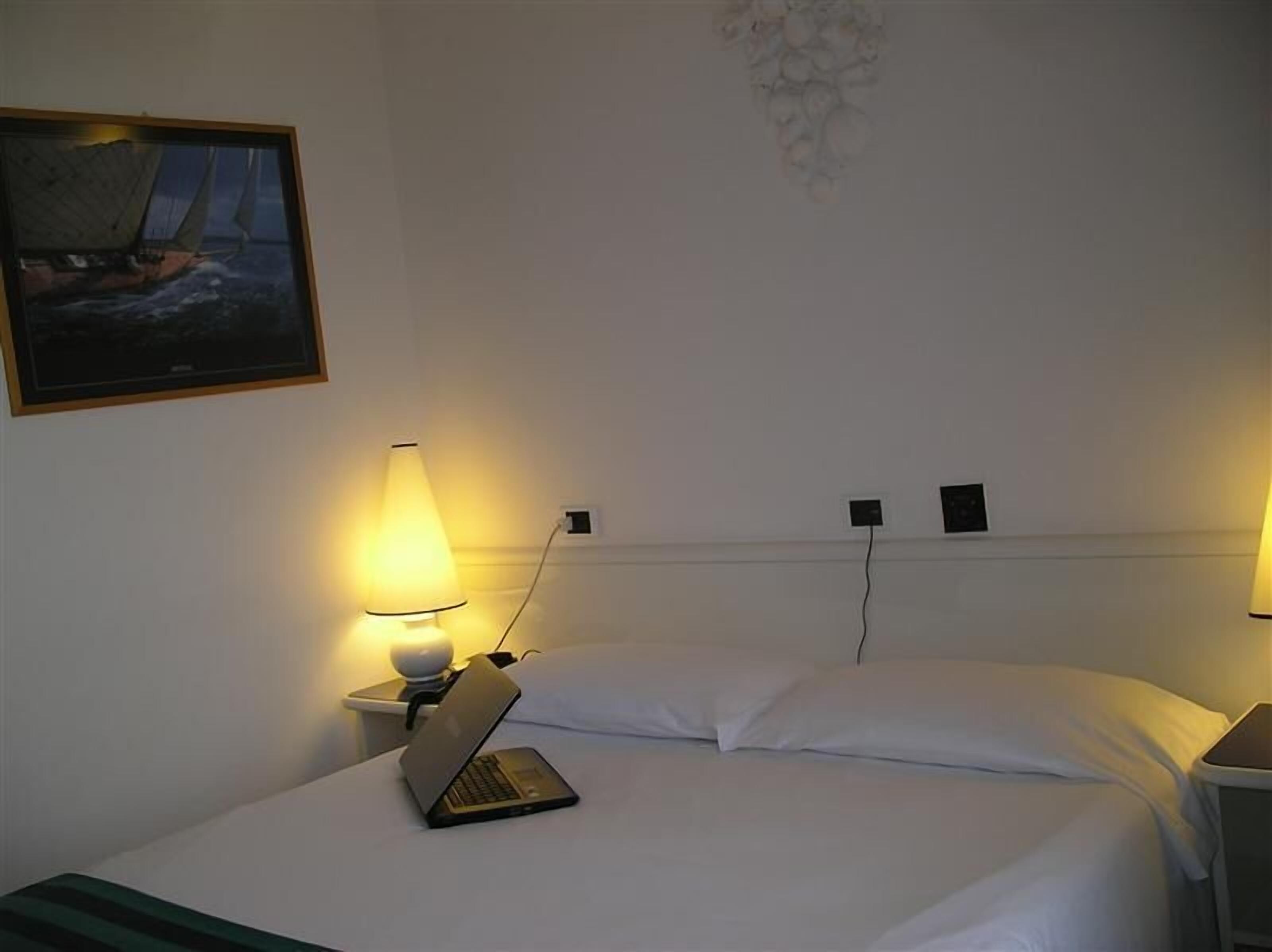Een hotelkamer met een bed, een laptop op het bed, een nachtkastje met een lamp en een schilderij aan de muur.