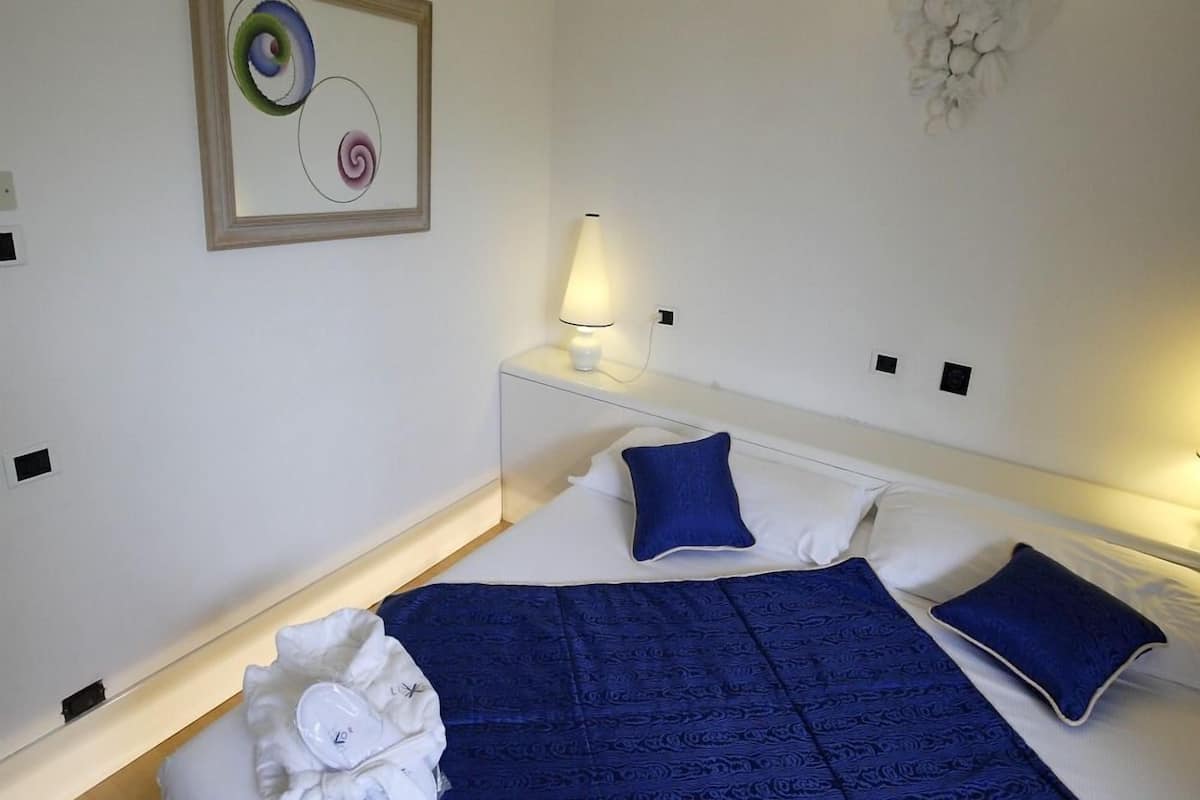 A hotel room with a large bed, a nightstand with a lamp, a wall-mounted light fixture, and a framed abstract painting.