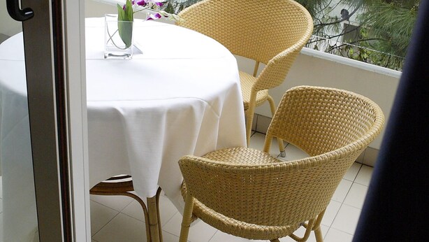Un balcon avec une table et deux chaises en osier, donnant sur un arbre et un immeuble.