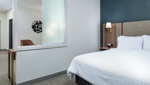 Quarto de hotel com uma cama grande, uma mesa de cabeceira, uma luminária na parede e uma janela com vista para outro quarto.