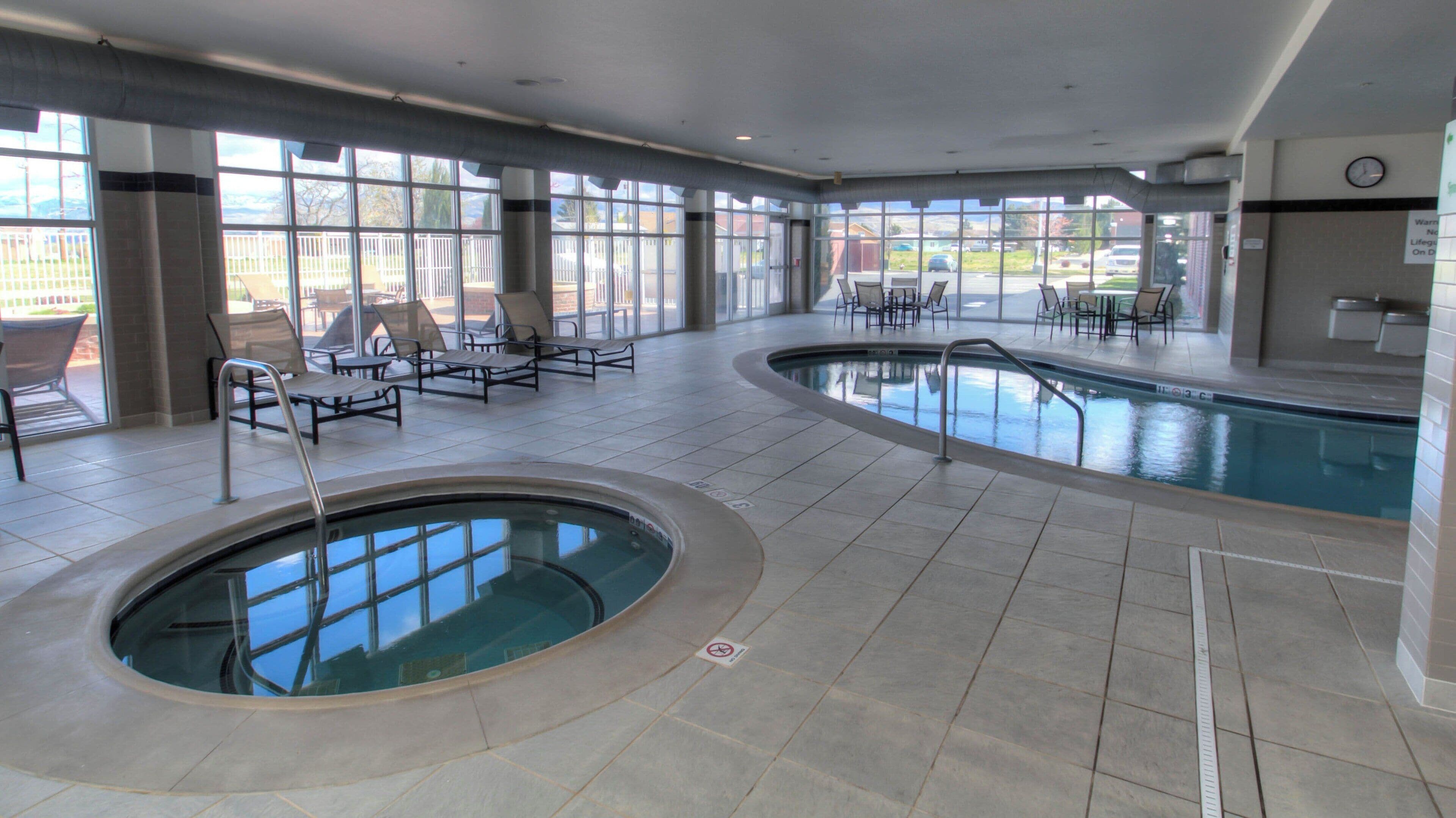 A large indoor swimming pool with lounge chairs and a sunbed, surrounded by large windows.