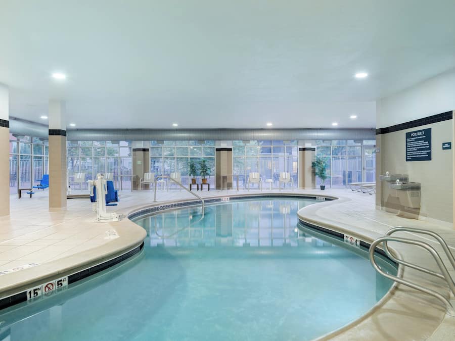 An indoor swimming pool with clear water, a pool lift, and a view of the outside through large windows.